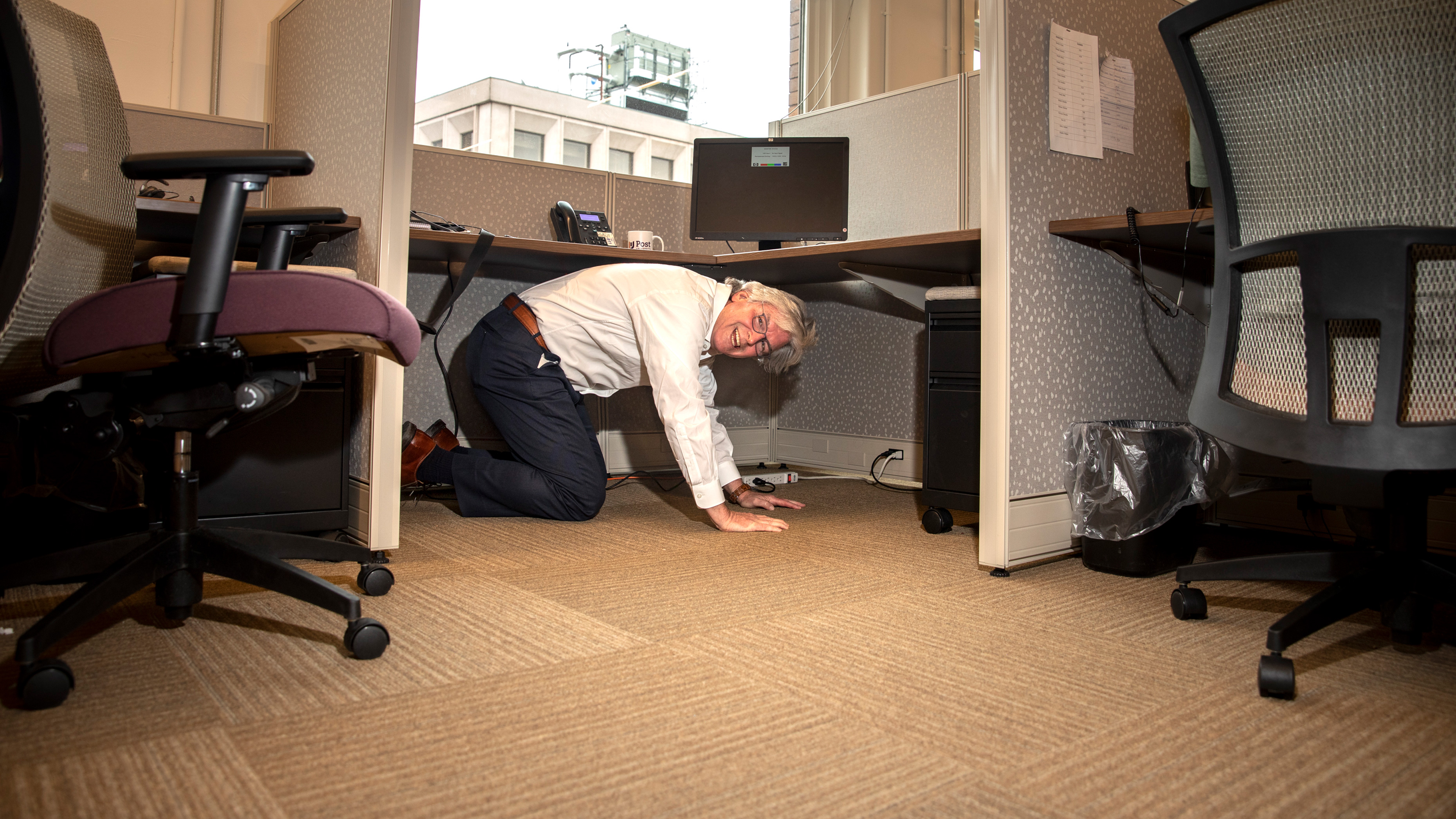 John Hopkins on hands and knees underneath an office desk and smiling