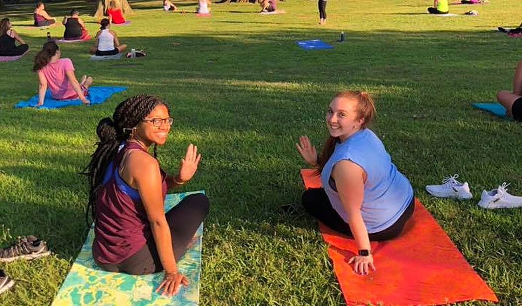 Two women sitting on yoga mats in the grass turned around and waving