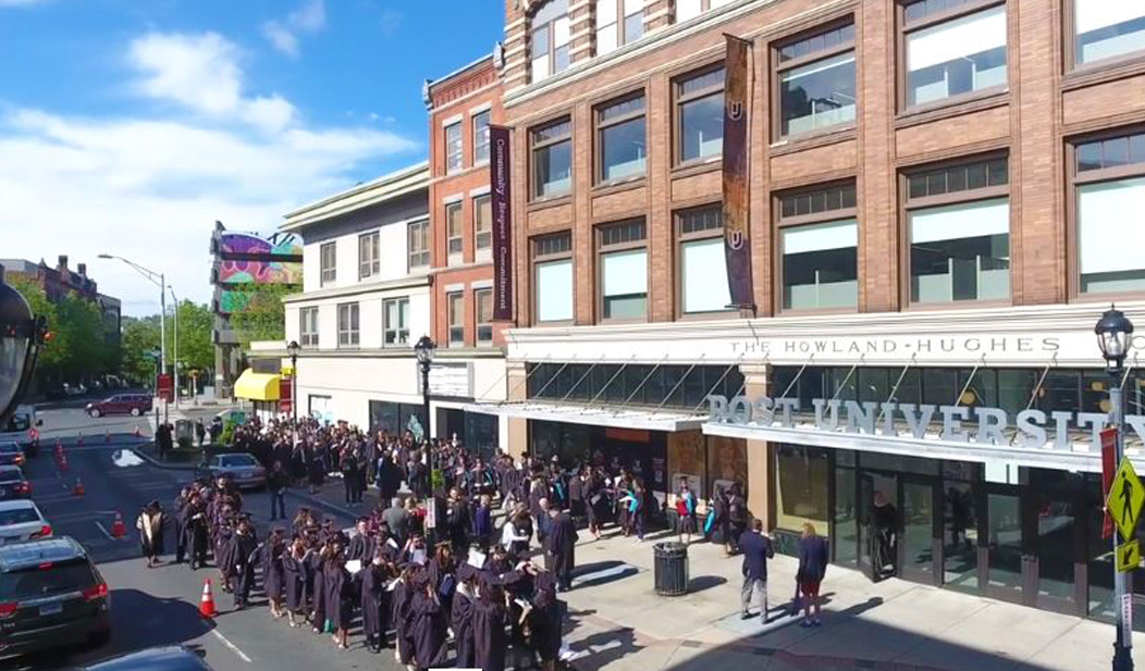 Graduates wearing caps and gowns lined up outside in front of Post University in Waterbury