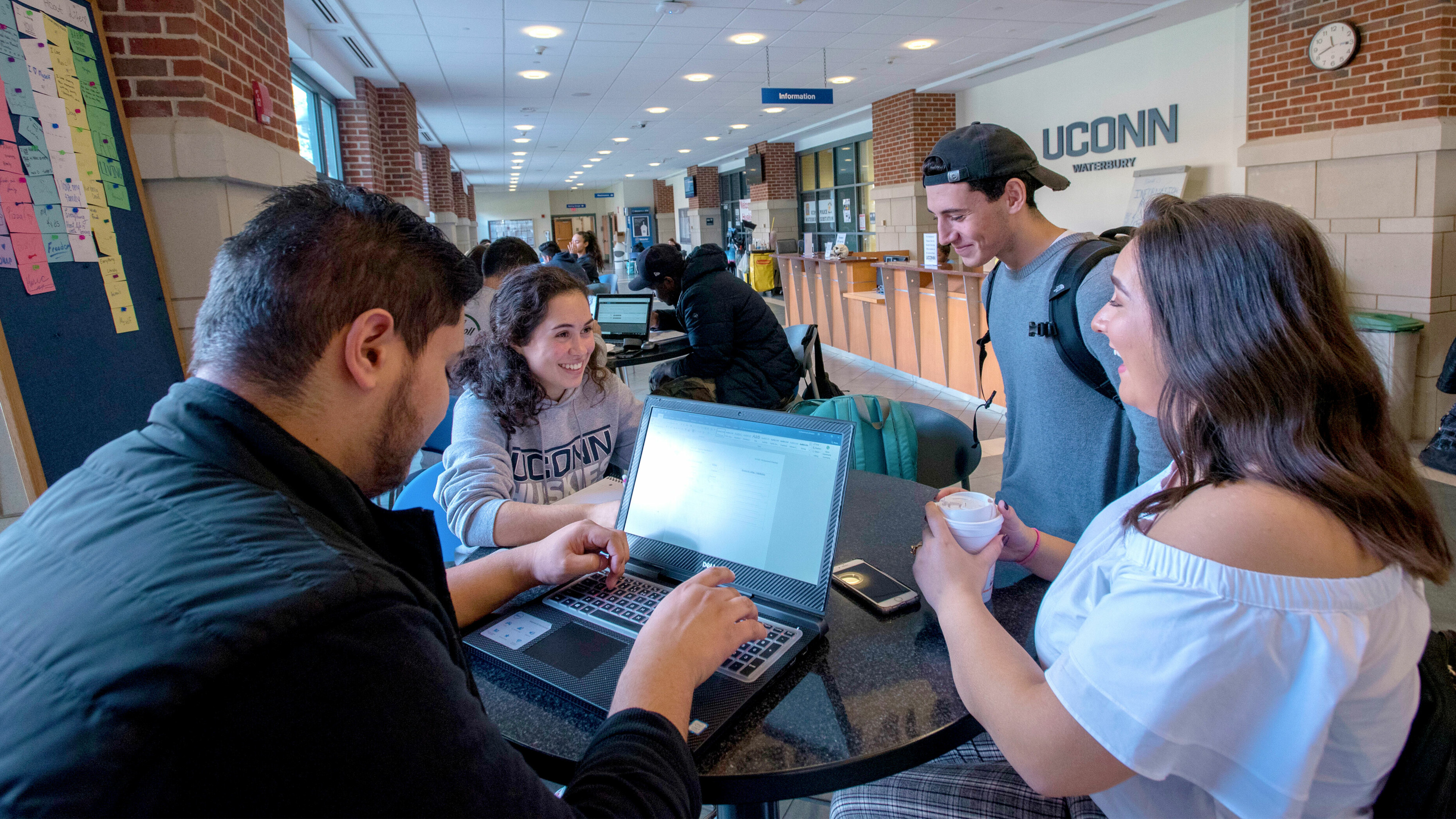 UConn Waterbury student working on laptop at a table with three other students having a conversation