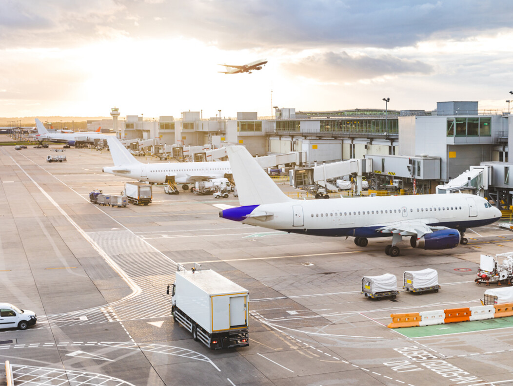 Airplanes and trucks parked outside of an airport with a plan taking off in the distance